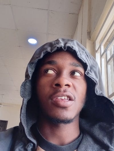 The image displays a close-up, head-and-shoulders portrait of a male individual positioned indoors. The subject has dark skin, a short dark beard on his chin, and is wearing a dark or grey hooded garment with the hood covering his head. A dark crew-neck shirt is visible beneath the hooded top. His eyes are directed upwards and to the viewer's left, while his mouth is slightly ajar, revealing his upper teeth. The background features a light-colored, possibly tiled, ceiling with a single illuminated circular recessed light fixture located above and to the left of the subject. To the subject's right, a light-colored wall or structural column is present, adjacent to a window secured with vertical metal bars. The scene is illuminated by artificial light. The image contains no visual information to identify a specific location within Kwazailewa, Nigeria.