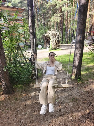 A young woman is sitting on a wooden swing in a forest setting. She is wearing a white tank top, a light-colored hooded jacket, and beige pants. Her right hand is raised to her mouth, and her hair is blowing in the wind. The swing is suspended between two large trees by ropes. In the background, there is a wooden structure made of sticks, a white sphere, and a multi-colored building. The ground is covered in pine needles and cones. The scene appears to be located in a rural or natural area, possibly on the outskirts of Onochino, Russia, given the vegetation and rustic structures.