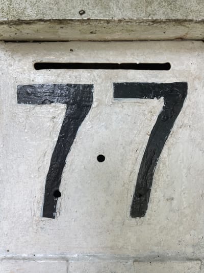 The image shows a close-up, eye-level, vertical shot of the number "77" painted in black on a weathered, off-white surface. The paint is thick and textured, with visible brushstrokes. The number "77" is composed of two large, bold digits. The left "7" is slightly more faded and textured than the right "7". There is a horizontal slit above the numbers, possibly a mail slot. Below the left "7" and slightly to the right, there is a small, dark circular hole. The surface appears to be part of an outdoor structure, possibly a mailbox or a building facade. The background above the numbers shows a rougher, discolored texture, suggesting age and exposure to elements. The overall impression is of an urban detail, likely a house or building number. Without further information, its specific location in Xuân Hương - Đà Lạt, Vietnam, cannot be determined.