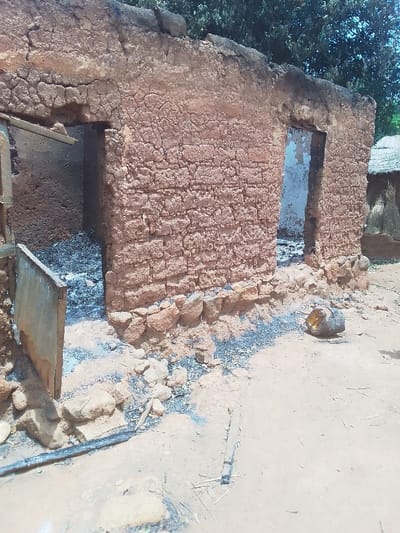 The image shows the remains of a mud brick building in Kwal, Nigeria (latitude: 9.804, longitude: 8.647). The building has two doorways. The left doorway is partially obscured by a wooden door, and the right doorway is open, revealing a charred interior. The building's exterior walls are severely damaged and cracked, with evidence of a fire. There are charred remains scattered on the ground in front of the building, and a large metal object lies on the ground near the right doorway. The ground is dry and dusty, and there are no people in the image. There is a single tree in the upper right corner of the image. The building is likely in a rural area, as the surrounding environment is open and undeveloped.