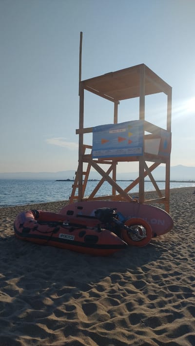 The image shows a beach scene in Neoi Epivates, Greece, with a wooden lifeguard tower in the background. In front of the tower, on the sand, are two inflatable watercraft and a paddleboard. The lifeguard tower is a tall, multi-level structure with a roof. A blue sign with various symbols and text in Greek is attached to the tower. The watercraft in the foreground is orange and appears to be a small inflatable boat with an outboard motor. Behind it is a pink paddleboard. The sea is calm and stretches to the horizon, with a faint outline of mountains in the distance. The sky is clear and blue, with the sun shining brightly. The sand is light brown and appears to be soft. The scene is set during the daytime, likely in the morning or late afternoon, given the angle of the sun and the shadows cast. There are no people visible in the image.