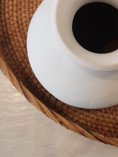 A close-up, overhead shot shows a white, round-bottomed vase with a wide opening. The interior of the vase is a deep, opaque black. The exterior of the vase has a matte finish with visible brushstrokes. The vase is placed on a woven, light brown wicker tray, which has a thick, twisted braided edge. The tray rests on a textured, light-colored surface, possibly wood or a similar material, with visible grain lines. The lighting suggests an indoor setting, with a soft, even illumination. No people are visible, and no specific time of day or weather conditions can be determined. The image is a still life, focusing on the textures and forms of the objects. There is no visible text. The scene appears to be set in Maitland, United States of America.