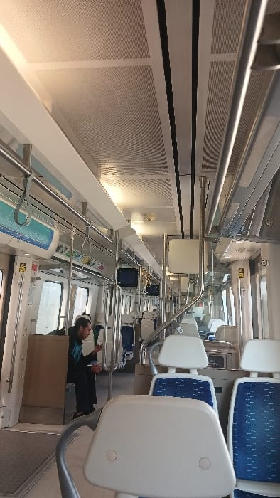 The image depicts the interior of a modern train car, likely within the Gaziemir district of Turkey. The perspective is from the front of the car, looking towards the rear. The ceiling consists of white, perforated acoustic panels interspersed with white beams and two parallel black strips running lengthwise, containing integrated lighting. Silver-colored grab poles and horizontal handrails with blue plastic loops are visible along the aisle and above the seats.

Seating is arranged in rows of individual seats. The seats feature white plastic shells for the back and headrests, with blue fabric inserts on the seat cushions, patterned with small white dots. Many seats are unoccupied, indicating a sparsely populated car.

On the left side of the aisle, approximately midway down the car, a single male individual is seated. He has dark hair, wears a dark jacket with light blue accents, and dark pants. He is looking down at a small handheld electronic device, presumably a mobile phone. Next to him, a light brown cardboard box rests on the floor.

The train interior appears clean and well-maintained. Windows line both sides of the car, offering views to the outside, which appears bright but indistinct. Several small, dark-screened monitors are mounted overhead along the length of the car. The floor is light grey, possibly carpeted or textured.