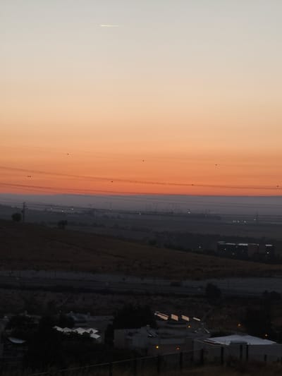 This is a vertical, eye-level shot of a landscape at dusk, likely taken from a higher vantage point overlooking a residential and industrial area. The sky transitions from a pale blue at the top to a vibrant orange hue towards the horizon, with a faint contrail from an aircraft visible in the upper portion. Several power lines crisscross the middle ground, some marked with small, dark circular insulators. Below the power lines, the landscape features rolling hills covered in dry, yellowish vegetation. In the distance, a cluster of industrial structures with tall cranes and lights is shrouded in a hazy atmosphere. Closer to the foreground, several low-rise buildings with angular, light-colored roofs are visible, some with illuminated windows. A dark metal fence runs along the bottom of the frame, separating the viewer's perspective from the scene below. The overall impression is one of a quiet, transitional moment between day and night in an Israeli settlement.