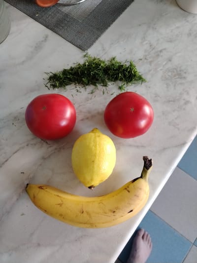 A close-up, high-angle shot shows a face made out of food items on a marble countertop. Two red tomatoes are positioned as eyes, a bright yellow lemon serves as the nose, and a curved banana forms the smiling mouth. A small pile of chopped green herbs is placed above the tomatoes, resembling eyebrows. The background is a kitchen countertop with a checkered mat in the upper left corner. The scene is located in the city of Rudzensk, Belarus.