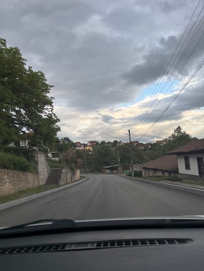 The image shows a street view in Baylovo, Bulgaria, captured from inside a vehicle. The road curves slightly to the right and leads uphill, with houses and trees lining both sides. A stone wall runs along the left side of the road. Power lines are visible against a cloudy sky with patches of blue. The interior of the vehicle, including part of a dashboard and air vent, is visible on the right side of the frame.