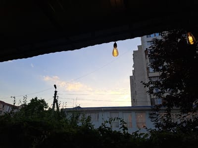 The image captures a view of a building and sky from under an overhang. The subjects are an architectural building, vegetation, and a clear sky with scattered clouds. An Edison-style light bulb hangs from a wire, illuminated and adding a vintage element to the scene. The building appears to be a multi-story structure with a light blue facade and white-framed windows on the lower portion. To the right, a taller building with a light-colored brick facade is visible. Dense green foliage, possibly a tree or bushes, occupies the foreground and parts of the mid-ground, creating a silhouette effect in some areas. A utility pole with wires is also present in the landscape. The time of day appears to be late afternoon or early evening, given the soft lighting and the colors of the sky, which range from pale blue to a soft yellow and pink near the horizon. The weather seems clear. The scene is likely an outdoor urban or suburban setting. There is no visible text in the image.