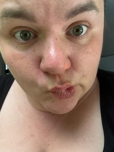 A close-up, forward-facing, unedited photo shows a person's face and upper chest. The person's skin is fair with visible pores and some freckles. The person has green eyes and light brown eyebrows. The person is wearing a black garment. The person has a small gold stud piercing on their right nostril. The person's lips are pursed, creating a pouty expression. The background is dark and out of focus. The image location is not identifiable from the provided visual information.