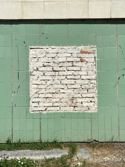The image depicts a section of an exterior wall in Krasnomakskoye Rural Settlement, Ukraine. The wall features a grid of light green square tiles. In the center, a rectangular section of these tiles has been removed, exposing a patch of white brickwork. The bricks are rough and appear weathered, with some red brick showing through. There are visible vertical lines between the tiles, indicating grout. The top of the wall is bordered by a band of light-colored, possibly stone or concrete, panels. The lower portion of the image shows a narrow strip of dry grass and some patches of green vegetation, with a concrete pathway visible in the foreground. There are some faint black spray-painted marks on the green tiles. No people are present in the image, and no actions are being depicted. The scene is static and appears to be an outdoor urban or suburban environment.