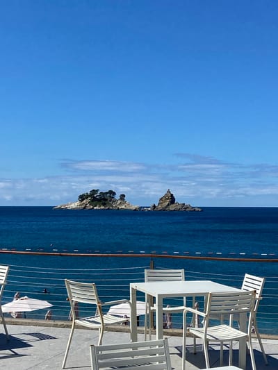 The image depicts a clear, bright day with a vast expanse of deep blue sea under a clear blue sky with scattered white clouds. In the distance, two rocky islands rise from the water. The larger island on the left is topped with lush green trees, while the island on the right is a jagged, imposing rock formation. In the foreground, a patio area is set up with white outdoor furniture, including several tables and chairs, positioned behind a railing overlooking the sea. A beach area with white umbrellas and beachgoers is visible below the patio. A person is swimming in the sea. The scene suggests a serene coastal setting.