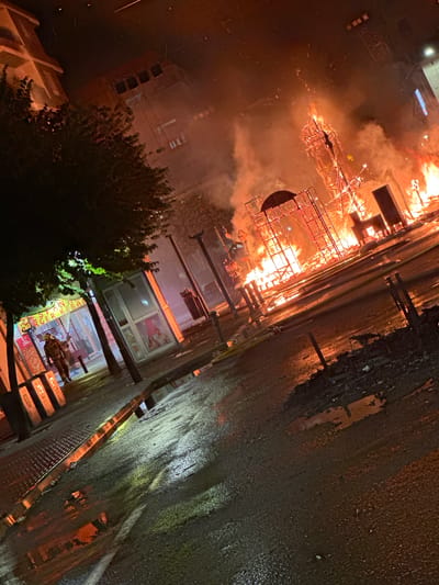 This image depicts a scene at night in Alicante, Spain, where a large structure, resembling a falla monument, is engulfed in flames. The fire is intense, with flames and smoke rising high into the air. The structure appears to be made of combustible materials and scaffolding, with parts of it collapsing or burning away. The surrounding environment shows signs of the event; a street or plaza is wet, reflecting the fire's glow. On the left side of the image, a tree shades a building with signage that appears to read "SPAIN BURGER KEBAB" in English and other languages. Two individuals, dressed in what appear to be firefighter uniforms, are walking away from the structure. In the background, buildings are visible, illuminated by the fire and smoke. The overall atmosphere is chaotic and dramatic, characterized by the destructive force of the fire.