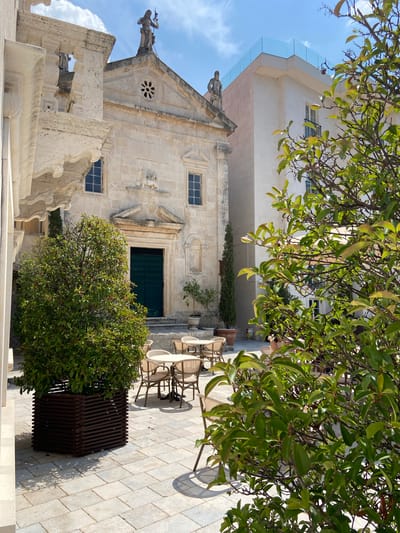 A sun-drenched square in Perast, Montenegro, features an ancient stone building with a green double door and a prominent pediment above. To its left, a balcony with intricate stonework juts out, and atop the building, two statues stand guard. Adjacent to this historic structure, a modern white building rises, crowned with a sleek glass railing. In the foreground, lush green trees partially obscure the view, their leaves catching the sunlight. The square itself is paved with light-colored stones, and several empty cafe tables and chairs are arranged, hinting at a leisurely atmosphere. The sky above is a bright blue, dotted with fluffy white clouds.