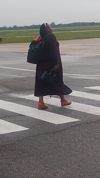 A full-shot, eye-level, outdoor, slightly blurry, vertical photograph shows a woman walking across a crosswalk on an asphalt road. The woman is wearing a dark purple dress, a black backpack, and pink wedge sandals. She is carrying a black purse in her right hand and looking up and to the right. The crosswalk has thick white stripes. In the background, a grassy field and trees are visible under an overcast sky. The scene is located at the Orerokpe Motor Park in Orerokpe, Nigeria.