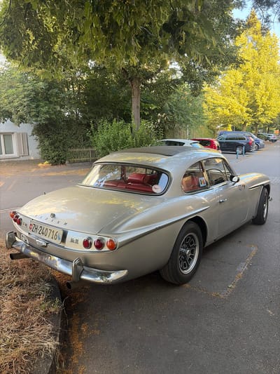 A silver vintage Jensen car, possibly a Mk III Interceptor, is parked on a paved street in Zurich, Switzerland. The car is a two-door coupe with a black sunroof and red leather interior visible through the windows. It has a chrome bumper and exhaust pipes. The license plate reads "ZH-240716". The car is parked next to a sidewalk with dry grass and leaves. In the background, there are trees with green and yellow foliage, a residential building with a window, and other parked cars. The sun is shining, casting shadows and highlighting the yellow leaves of a tree on the right side of the image. The overall scene suggests a sunny day in a residential area.