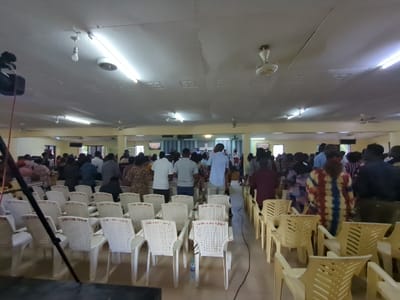 The image shows the interior of a church, likely in Morogoro, Tanzania. The coordinates provided (-6.829, 37.658) locate the city of Morogoro. The scene depicts a congregation of approximately 50 people standing in a large, rectangular room with a tiled floor. The room is sparsely furnished, with rows of yellow plastic chairs facing a stage at the back. The stage is equipped with a screen displaying an unknown message. Most of the people are dressed in casual attire, and some are wearing hats. The ceiling features several fluorescent lights and a ceiling fan. A large black microphone stand is positioned at the bottom of the frame. Several people are standing near the stage, apparently engaged in a religious service or prayer. The overall atmosphere seems peaceful and serene.