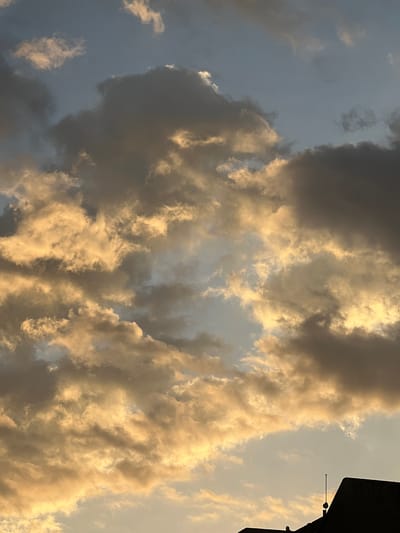 The image shows a sky filled with dramatic clouds illuminated by the warm, golden light of either sunrise or sunset. The clouds are a mix of dark grays and bright yellows, creating a textured and dynamic sky. The lower portion of the image features the silhouette of a building with a dark, angular roofline and a thin antenna. The setting is likely urban, given the building, and the lighting suggests it is either early morning or late evening. The specific location in Maracay, Venezuela, is not discernable from the image itself, as there are no identifying landmarks or contextual clues.
