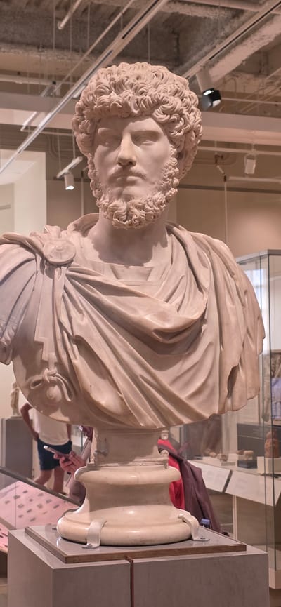 The image shows a marble bust of a man with curly hair and a beard, likely a historical figure from Roman times, displayed on a pedestal in a museum. The bust is made of light-colored marble and is intricately carved, depicting realistic facial features and drapery. The man is wearing a toga-like garment secured with a brooch on his shoulder. The background suggests an indoor exhibition space with display cases, lighting fixtures on the ceiling, and other museum visitors in the distance. One visitor in the mid-ground is holding a smartphone. The overall lighting is consistent with museum interior lighting.