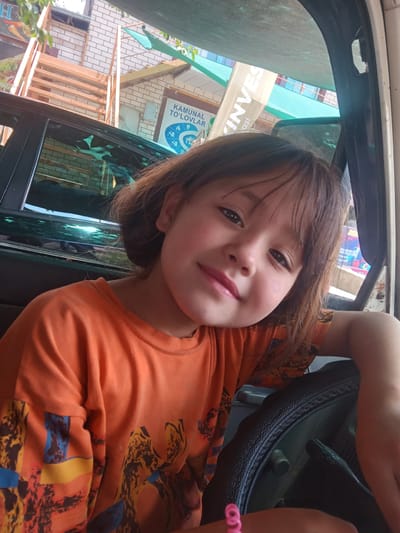 A young girl with brown hair and dark eyes smiles at the camera, leaning against a steering wheel inside a vehicle. She is wearing an orange t-shirt with a colorful, abstract pattern. Her left arm is bent, resting on the steering wheel, and her right arm is visible on the right side of the frame. The interior of the vehicle appears to be old or worn, with a grey dashboard. Outside the car, through the windows, a building with a brick facade and light-colored horizontal siding is visible. A banner with text, partially obscured and rotated, hangs on the building. Green foliage is also visible. The lighting suggests it is daytime. The text on the banner appears to be in a non-Latin script and includes the word "INVEST". Another sign with blue and white text and icons is visible. The scene appears to be in Obizarang, Uzbekistan, based on the location context. The girl's expression is happy and playful, suggesting a casual or fun moment captured inside the car.