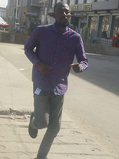 The image displays a male individual in motion on a paved surface in an urban area of Babban Giji, Nigeria. The subject, a man of African descent with a beard, is captured mid-stride, facing slightly right of the camera. He wears a long-sleeved blue and red plaid button-up shirt and dark trousers, from which a white tag or label is suspended near his left hip. His left leg is elevated, suggesting movement, and his right arm is bent with a closed fist. The environment consists of a light-colored sidewalk in the foreground, transitioning to a darker paved road. In the background, a commercial building features glass windows and illuminated signage, partially reading "FRESHER. BETTER." and "ENTRANCE." Additional background elements include a pillar, a partially visible sign displaying "24," and a red awning-like structure on the far left. The scene is under bright daylight, indicated by strong illumination and faint shadows.
