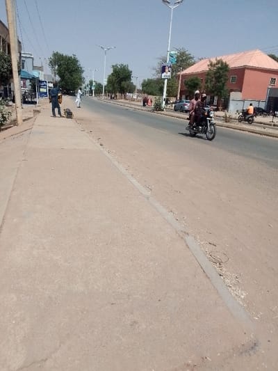 A sunlit street scene shows a two-lane road with a wide, sandy shoulder and a concrete sidewalk. Several people are visible, including two men on a motorcycle, a man standing on the sidewalk, and a few others in the distance. Buildings, trees, and streetlights line the road. A campaign poster can be seen on a pole. The sky is clear.