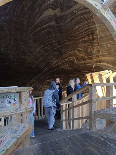 A group of people are standing on a wooden walkway inside a cave. The cave walls are made of salt, with distinct layers of brown and white minerals. The walkway is made of rough-hewn wood, with benches and railings. Some of the wood is covered in graffiti.
In the foreground, a wooden bench is visible, covered in colorful graffiti. Behind it, a young person wearing a pink hoodie and jeans stands on the walkway, looking to the side. Further back, a man with a beard and a baby on his chest is visible. He is wearing a grey hoodie. To his right, two women are standing. One woman, wearing glasses and a denim jacket, is taking a photo with her phone. The other woman is wearing a black jacket.
The cave ceiling is high and arched, with visible geological formations. A netting-like material is hanging in some sections of the cave. The lighting in the cave appears to be artificial, casting a warm glow on the wooden structures. The overall impression is of a tourist attraction where visitors are exploring the unique salt formations.
Visible text includes "ANDRA", "Les kangowed 25", "PL", "Mokus", "FA", "ZSOLTO 2034", "FIGI DARDAC", "DANCA", "FIPR", "2Rok You", "ANAD", "MIRASJONEL", "GABOR ALIN", "KITTY", "Andina", "N", and "Davel". The location is identified as Turda, Romania. The time of day and weather are not discernible from the image. The emotions of the people are not clearly visible, but they appear to be engaged in observing their surroundings.