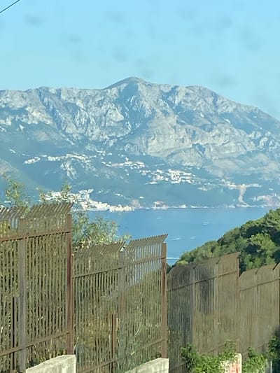 In Seoce, Montenegro, a view is captured overlooking a bay with a mountainous backdrop. In the foreground, a weathered metal fence, composed of vertical bars, runs horizontally across the frame. Beyond the fence, lush green trees and vegetation cover a hillside that slopes down towards the water. The bay itself is a calm expanse of blue water, with distant white buildings scattered along the coastline at the base of the mountains. These buildings appear to be nestled into the terrain. The mountains in the background are rugged and rocky, with clear ridges and valleys, under a bright, pale blue sky. There are no people visible in the image.