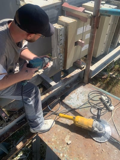 A man wearing a black baseball cap, a grey t-shirt, and grey work pants is using a teal cordless drill to work on a large, tan-colored electrical component. He has a silver chain around his neck and a tattoo of an airplane on his left forearm. He is standing on a rusty metal platform, which appears to be part of an industrial structure. On the platform next to him are a yellow angle grinder with a cutting disc, coiled black electrical cords, and a power strip with multiple outlets. The background shows a grassy area with some scattered debris, suggesting an outdoor, possibly industrial or construction site. The overall scene is outdoors under bright sunlight. The image was captured in Zelena Polyana, Ukraine.