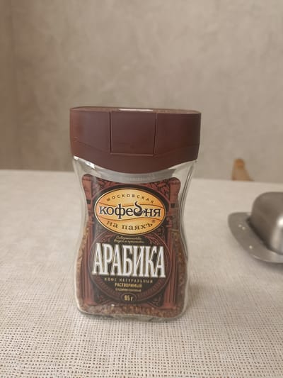 The image shows a glass jar of "Arabika" instant coffee, which appears to be a Russian brand called "Moskovskaya Kofeynya." The jar is brown and transparent, filled with coffee granules. The label on the jar features the brand name prominently, along with text in Russian indicating "coffee on wax," "perfection of taste and aroma," "natural instant coffee," and "freeze-dried." The net weight is stated as "95g." The jar is placed on a white, textured tablecloth. In the background, a plain, light-colored wall is visible. To the right of the jar, a portion of a shiny metal object, possibly a butter dish or lid, is present. There are no people visible in the image. The scene is indoors, with neutral lighting. The location is not specified, and there is no indication it is in the city of Izberbash, Russia.