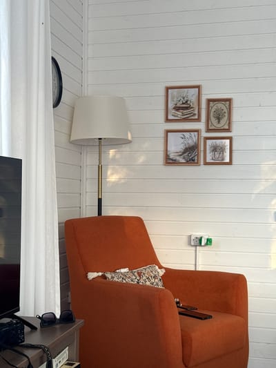 The image depicts an indoor scene, likely a living area, in the village of Starokorsunskaya, Russia. A bright orange armchair is positioned in the foreground, slightly right of center. A small decorative pillow with a floral pattern rests on the armrest of the chair. Two smartphones are placed on the seat of the armchair. To the left of the armchair, a dark brown side table holds a pair of black sunglasses, a camera, and other unidentifiable items. A white sheer curtain hangs on the left side of the frame, partially obscuring a black-faced clock on a white paneled wall. A tall floor lamp with a cream-colored lampshade stands behind the armchair. The wall is made of white horizontal planks, and four small framed pictures are arranged on it above the armchair. The pictures feature botanical and landscape illustrations. A white electrical outlet with a green adapter is mounted on the wall to the right of the armchair. The lighting suggests it is daytime, with sunlight casting shadows on the wall.