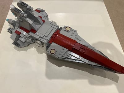 A grey and dark red LEGO model of a spacecraft, resembling a starship, rests diagonally on a smooth, light-colored surface. The model is oriented with its multi-engine stern section towards the upper-left and its tapering, pointed bow towards the lower-right of the frame. The primary color scheme consists of light grey bricks forming the hull and structural elements, accented by dark red plates and bricks along the central spine and sides. Transparent light blue circular elements are visible at the rear, along with transparent clear elements atop some fin structures. Subtle shadows are cast by the model onto the surface, indicating an overhead light source. No individuals are present. This scene is located in Abbotsbury, United Kingdom.