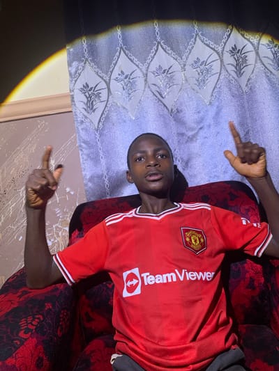 A young boy is sitting on a red and black patterned armchair in Barakin Sabawa, Nigeria. He is wearing a red Manchester United jersey with white trim. The boy is looking at the camera and has his index fingers pointed upwards. The background consists of a light-colored curtain with a decorative pattern and a wall with splattered paint. A bright light source casts a distinct shadow on the left side of the image.