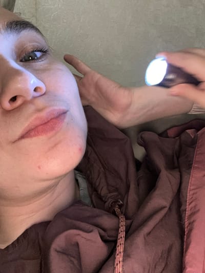 An image from a low angle shows a fair-skinned person lying on what appears to be a light-colored floor. The person has long eyelashes, and their face is angled towards the camera. Their lips are colored, potentially with lipstick, and there is a reddish blemish on their cheek. A hand holding a flashlight is positioned above the person's head, illuminating their face. Part of a dark-colored, possibly maroon, fabric or garment is draped to the side of the person. The image was taken from a tilted angle. No details confirm a location in the city of Uva, Russia.
