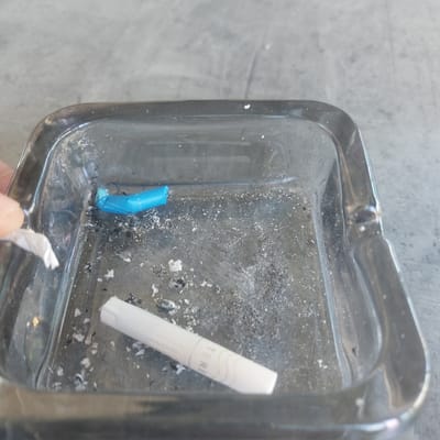 The image shows a close-up of an ashtray containing cigarette ash and two cigarette butts. One butt is white with some green lettering visible, and the other is a blue filter. There is also a small piece of white paper in the ashtray, and a hand is partially visible on the left side, holding another small piece of paper. The ashtray is made of clear glass and sits on a textured gray surface. The scene appears to be outdoors, as indicated by the lighting and the visible background. There are no specific landmarks or details in the image that would allow for pinpointing a location in Ablanitsa, Bulgaria.