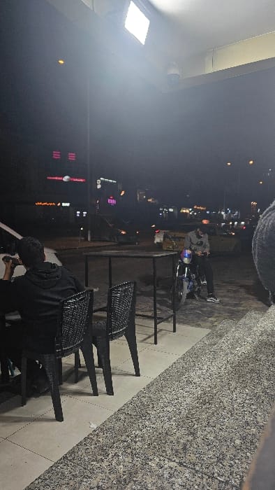Night scene shows a street with sparse city lights. A person is on a motorcycle next to outdoor cafe furniture, including chairs and tables. The outdoor area appears to be a terrace or patio with a stone or tile wall. There is a strong light source on the ceiling illuminating the area. The overall image is dark with light reflecting off surfaces.