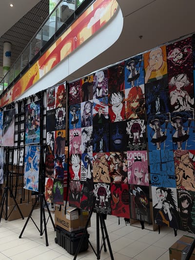 This image depicts an indoor display of anime-style posters. The posters are hung on a metal rack system and feature various characters from anime series. Some of the recognizable characters include Monkey D. Luffy from One Piece, and characters from Death Note. The posters are arranged in a grid pattern. In the foreground, there are several black tripods supporting some of the posters, and some cardboard boxes and a black rolling case are visible on the tiled floor. In the background, there is a railing with a decorative banner above it. The environment appears to be a public indoor space, possibly a mall or convention center. There are no people clearly visible in the image. The overall scene is a commercial display of anime merchandise. This image is not related to Gorzow, Poland.