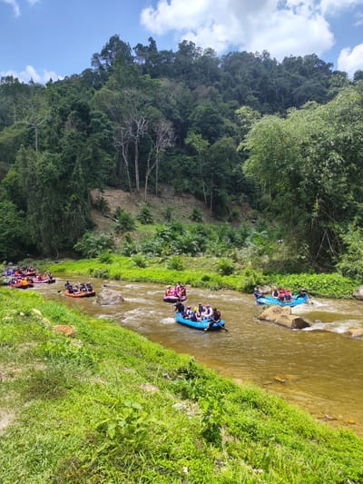 The image depicts a group of people participating in a white water rafting activity on a river in Tambon Song Phraek, Thailand. Several inflatable rafts, each carrying multiple individuals, are navigating the river's current. The river water is a murky brown, and rocks are visible both within and along the riverbanks. Lush greenery, including various trees and vegetation, covers the surrounding landscape and rises up the slopes in the background. The sky is blue with some white clouds.