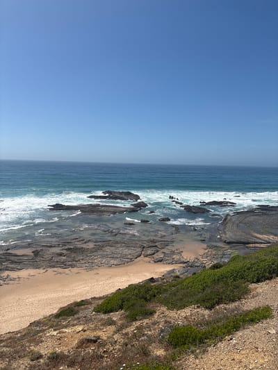 This image depicts a sunlit coastal landscape near Aljezur, Portugal, likely along the Costa Vicentina. The scene is dominated by a clear, azure sky occupying the top third of the frame. Below, the Atlantic Ocean stretches towards the horizon, displaying varying shades of blue. Waves crash against the shore, creating white foam and turbulent water around numerous dark, irregularly shaped rock formations that protrude from the water.

Closer to the viewer, the shoreline reveals an intricate geological structure of layered, dark grey rock formations interspersed with patches of light brown sand. A small, crescent-shaped sandy beach is visible in the lower left quadrant. The immediate foreground and right side of the image feature a sloping cliffside or hillside covered with vibrant green low-lying shrubs, sparse dry vegetation, and exposed reddish-brown earth and small stones. No people, man-made structures, or animals are visible in the photograph. The overall impression is one of a rugged, natural coastline under bright daylight conditions.