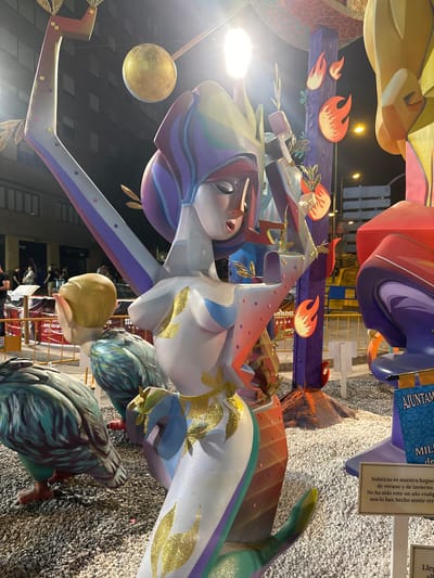 The image shows a nighttime outdoor scene featuring elaborate, colorful sculptures that appear to be part of a festival or parade. The sculptures are made of a material resembling papier-mâché or painted wood and depict stylized figures and decorative elements. In the foreground, a large sculpture of a woman with a flowing headdress and a white, glittery garment adorned with gold leaf details is prominent. To the left and slightly behind her are two large, feathered bird-like sculptures. The background includes other large, abstract sculptures in bright reds, purples, and yellows, as well as illuminated streetlights and buildings. A street sign with the text "AJUNTAMIENTO DE ALICANTE" is visible in the lower right corner, indicating the location is Alicante, Spain. The ground is covered in white gravel. Several people are visible in the background, suggesting the presence of an event with attendees. The overall impression is festive and artistic, likely related to a local celebration in Alicante.