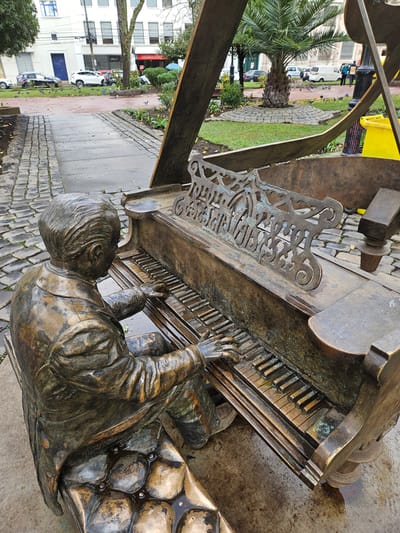 The image depicts a bronze sculpture of a man playing a piano in an outdoor urban setting. The sculpture is detailed, showing the man's attire, his hands on the piano keys, and the piano itself, which appears weathered. The piano has an ornate metal sheet music stand. The ground around the sculpture is paved with cobblestones and appears wet, suggesting recent rain. In the background, buildings and trees are visible, along with parked cars and a few pigeons. To the right, a yellow trash can and a lamppost are present. This sculpture is located in Chillán, Chile, specifically in Plaza San Juan Bosco, a park within the city.