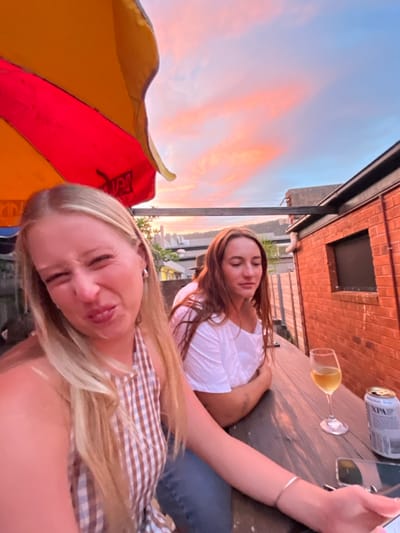 Two young adult females are depicted on an outdoor patio or rooftop venue in Thirroul, Australia, under a vibrant sunset sky. The woman in the foreground, positioned to the left, has long blonde hair and is making an exaggerated, grimacing expression with squinted eyes and pursed lips. She wears a brown and white checkered halter top. Her left arm is visible in the lower right, holding a smartphone. A thin silver bracelet adorns her left wrist.

To her right, a second woman with long brown hair, wearing a white off-the-shoulder top and blue jeans, is seated or leaning against a dark wooden surface. Her expression is neutral, with her gaze directed slightly downwards. On the wooden surface in front of her are a wine glass containing a light amber liquid and a silver beverage can partially visible, labeled "XPA."

Above the women and to the left, a portion of a red and yellow striped patio umbrella is visible. To the right, a red brick building with a dark-framed window is present, topped by a dark metal beam structure. The background features a dramatic sunset sky with soft pink, orange, and light blue hues, indicating either sunrise or sunset. A distant landscape of hills and urban structures is visible along the horizon beneath the colorful sky. The overall lighting is warm, emanating from the setting sun.