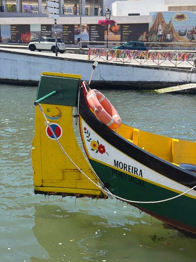 This image shows a close-up of the stern of a traditional Moliceiro boat docked in the city of Aveiro, Portugal. The boat has a yellow stern with green trim, and is decorated with floral motifs and the name "MOREIRA PORTUGAL" written in black on its green hull. Two orange life preservers are attached to the stern. The boat is floating in murky green water, with reflections of the boat and its surroundings visible on the surface. In the background, a white car is parked on a street bordering a canal, with a building behind it. The street is lined with posters and colorful ribbons are draped along a railing. The scene suggests a sunny day in a canal-side area of Aveiro, known for its Moliceiro boats and waterways.
