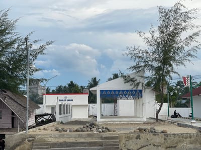 In Lancok, Indonesia, a public toilet facility stands adjacent to a dilapidated wooden structure with a thatched roof. The toilet building is modern, painted white with a red trim and features blue decorative panels above an open-air entrance. A paved path with white posts and black and yellow striping runs in front of the facility, where two people are seen riding motorcycles. A slender tree with sparse foliage grows in front of the modern building. The foreground is a rough concrete embankment scattered with large, smooth stones. The sky above is filled with a mix of white and gray clouds.