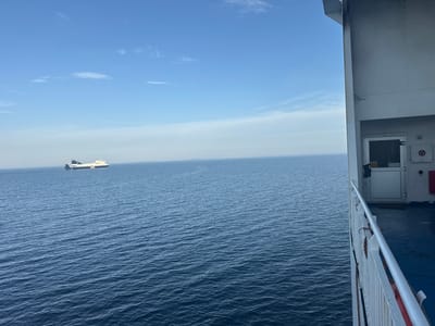 A ferry is seen sailing on a calm, blue ocean under a clear sky with sparse clouds. The perspective is from another vessel, with the railing and part of its superstructure visible in the foreground on the right. The ferry is white with blue and black accents, and appears to be a roll-on/roll-off ferry. The water is dark blue and shows gentle ripples. In the far distance, a faint outline of land is visible on the horizon. The scene is likely taking place in the waters off Sweden, given the text "UPOS" visible on the side of the ferry, which is a known ferry operator in the region.
