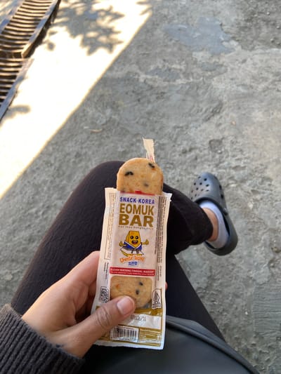 A person, viewed from above, sits on a paved surface. They are wearing dark-colored pants and a grey long-sleeved shirt. Their left hand is holding a snack bar with a white and yellow wrapper. The snack bar, labeled "SNACK KOREA EOMUK BAR," features a cartoon character and text in Indonesian and Korean. The snack itself is visible at the top of the wrapper, appearing to be a light-colored bar with dark specks. To the person's right, their right leg is visible, wearing a dark grey Croc shoe over a white sock. The ground is rough and grey, with some areas appearing lighter due to sunlight and shadow. A metal grate with horizontal bars is visible in the upper left corner of the image. The scene suggests a casual outdoor setting, possibly a park or a street in Margorejo, Indonesia, given the presence of Indonesian text on the snack wrapper.