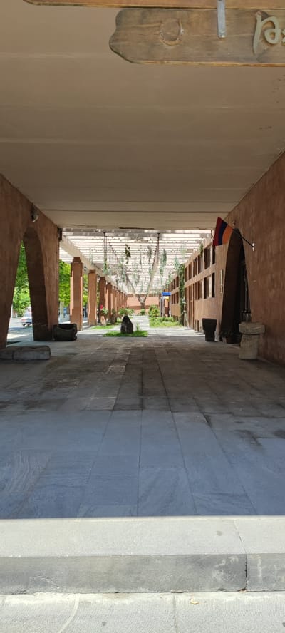 The image displays a long, covered pedestrian walkway in Vagharshapat, Armenia, extending into the distance under bright daylight. The perspective is from a slightly elevated point, looking directly down the central axis of the walkway.

The overhead structure features a flat, light beige ceiling. A dark brown wooden sign, attached by a metal bracket, hangs from the ceiling in the upper center. The sign bears a light-colored horseshoe symbol on its left and Armenian script reading "ԲԱՐԻ" (Bari) on its right.

The left side of the walkway is defined by a wall of reddish-brown brick or stone, incorporating large, rounded arches. Through the leftmost arch, a paved street is visible, along with the rear portion of a white sedan parked on the street. A dark, squat stone object rests on the ground near the base of this arch.

The right side features a solid reddish-brown wall. An Armenian national flag, composed of horizontal red, blue, and orange stripes, is mounted to this wall and extends diagonally into the walkway. Further into the scene on the right, another arched opening is partially visible.

The ground of the walkway is paved with large, rectangular grey tiles. The immediate foreground shows a light grey stone step or curb with distinct concentric circular patterns on its surface, leading up to the main walkway level.

Beyond the initial covered section, the walkway opens into a brighter area. A trellis system, adorned with numerous small green plants suspended from it, forms a green canopy overhead. This trellis structure is supported by a series of square, reddish-brown pillars that line both sides of the path, extending into the background.

The paved path in this more open section is bordered by small, irregular patches of green grass. Several large, dark, naturally shaped stones or sculptural elements are placed within these grassy areas, with one prominent dark stone centrally located on the left. Other similar stone elements are visible along the right side of the path further back. The distant end of the walkway transitions into an outdoor landscape with visible trees and greenery, illuminated by strong sunlight that casts distinct shadows along the walkway. No individuals are present in the scene.
