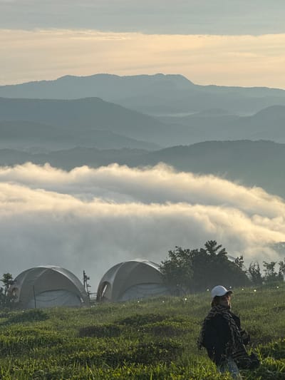 In the early morning light, a person wearing a white baseball cap, sunglasses, and a patterned scarf stands in a grassy field. Behind them, two dome-shaped tents are pitched on a hillside. Beyond the tents, layers of mist-covered mountains recede into the distance. The sky is a soft gradient of pale yellow and blue, with a few wispy clouds. The overall scene suggests a serene camping or glamping experience in a mountainous region, likely in the city of Phát Chi, Vietnam, known for its beautiful landscapes and misty mountains.