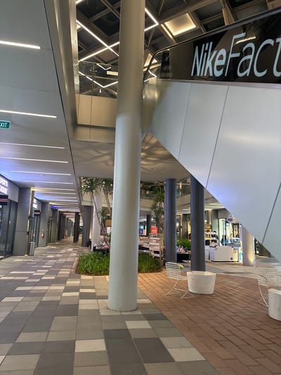 The image depicts an outdoor corridor at a shopping center in Wanasari, Indonesia. The corridor is lined with shops on one side and a modern, architectural walkway on the other, featuring a large, curved white canopy overhead. The walkway is illuminated by linear ceiling lights. Large, cylindrical pillars support the canopy. The floor is paved with rectangular bricks in various shades of brown and grey, creating a checkered pattern. There are also sections of grey square tiles. White metal chairs and planters with green foliage are placed along the walkway, suggesting an area for relaxation or waiting.

In the distance, shops are visible, with some displaying merchandise and signage. One prominent sign reads "Nike Factory" in white lettering against a black background. Other smaller signs are also visible, some with text that is difficult to discern due to the image quality and distance.

There are a few people visible in the distance, appearing to be shoppers or employees within the shops or moving through the corridor. The overall atmosphere seems to be that of a contemporary commercial space, possibly in the late afternoon or early evening given the artificial lighting. The weather appears to be clear. The scene is clean and well-maintained, indicative of a retail environment designed for a pleasant shopping experience.