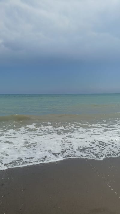 The image depicts a serene beach scene with a calm sea under a partly cloudy sky. The foreground shows dark sand wet from the receding tide, with a line of white foam marking the edge of the water. Gentle waves are breaking on the shore, creating a frothy texture. The sea itself is a tranquil turquoise, stretching to the horizon where it meets a pale blue sky. A band of soft, diffused clouds occupies the upper portion of the sky, obscuring direct sunlight and lending a muted quality to the scene. The overall impression is one of peacefulness and natural beauty. This scene is consistent with many beaches found along the coastline of Mezitli, Turkey, known for its Mediterranean shores and clear waters.