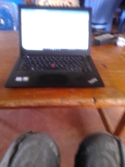 The scene depicts a black laptop computer resting on a light brown wooden table, observed from a seated perspective in Ndeiya, Kenya. The laptop screen is illuminated, displaying a predominantly white background with a taskbar and several application icons visible at the bottom. A red trackpoint is present on the black keyboard. Below the table, the knees of an individual, clad in dark fabric, are partially visible. In the blurred background to the left of the laptop, a blue object with a ribbed texture is present. To the right, out-of-focus items include a gray rectangular power adapter or device and crumpled white material, with a blue vertically striped pattern visible further back.