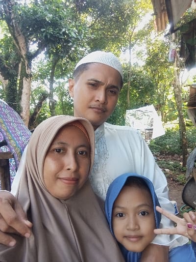 This image displays three individuals – one adult male, one adult female, and one young female child – posing outdoors in Bulukandang, Indonesia. The adult male is centrally positioned, wearing a white peci and a white embroidered long-sleeved shirt. The adult female is in the lower-left foreground, wearing a light brown hijab and matching attire, gazing towards the camera. The young female child is in the lower-right foreground, wearing a blue hijab and blue clothing, extending her left hand in a "peace" sign with pink-painted fingernails. The background consists of dense green foliage and tree trunks, indicating a natural, possibly rural or semi-urban, setting with bright, filtered sunlight. Visible structural elements include a patterned fabric on the left and a partial wooden structure with hanging objects on the upper right.